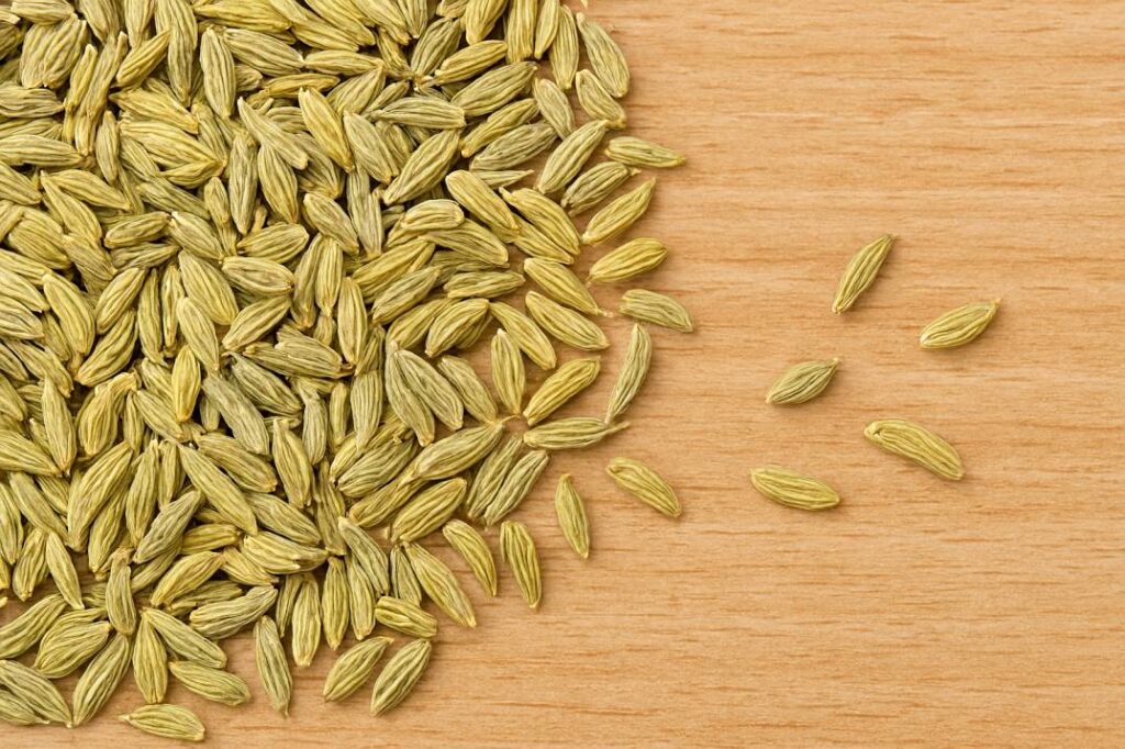 A close-up view of fennel seeds scattered across a light wooden surface. The seeds are elongated, slightly curved, and ridged, with a pale greenish-brown color. Some seeds are grouped together while others are spread out individually, revealing the wood grain beneath.